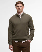 Man wearing a green sweater over a plaid shirt with brown pants on a light gray background