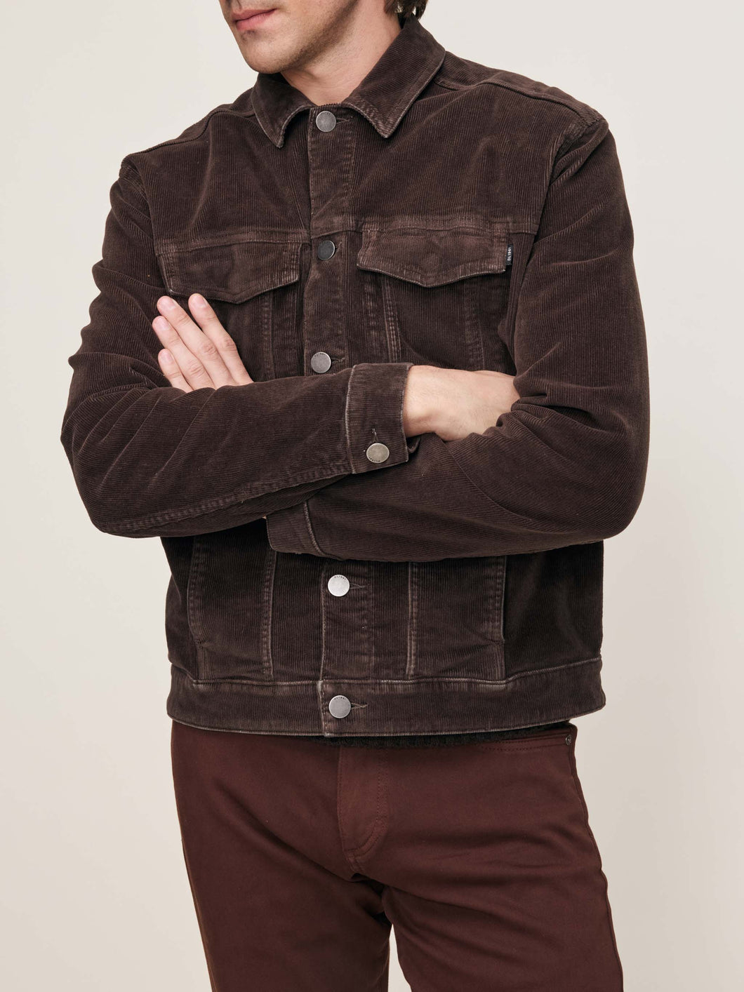 Man wearing a brown corduroy jacket with arms crossed on a beige background
