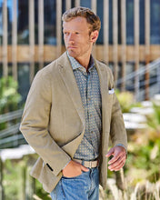 Man wearing a beige blazer over a patterned shirt with blue jeans, standing outdoors.