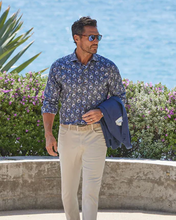 Man in a blue patterned shirt and beige pants standing by a body of water with greenery.