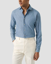 Man wearing a blue button-up shirt and white pants on a light gray background