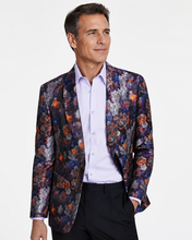A man wearing a dark floral patterned blazer over a light purple dress shirt and black trousers, photographed against a plain light gray studio background.