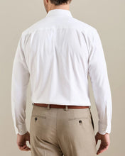 A man viewed from behind wearing a white long-sleeve dress shirt tucked into beige trousers with a brown belt, against a plain beige studio background.