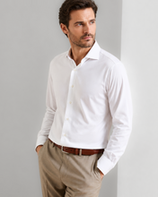 A man wearing a white long-sleeve button-down dress shirt, khaki trousers, and a brown leather belt, standing against a light gray wall.