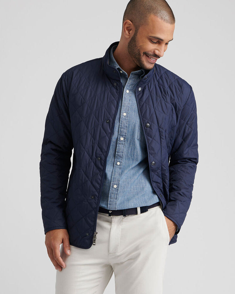 Man wearing a navy quilted jacket over a blue shirt with white pants on a light gray background