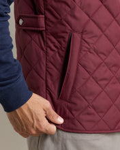 Close-up of a person wearing a quilted burgundy vest over a navy blue long-sleeve shirt, with a hand resting near the hem.