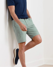 Man wearing a navy polo shirt and light green shorts against a white wall.