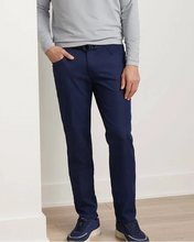 A person wearing slim-fit navy blue trousers, a light gray long-sleeve shirt, and navy sneakers, standing in a minimalist room with white walls and light wood flooring.
