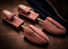 Three wooden shoe tree inserts with brass buckles arranged on a dark wooden surface next to a brown leather shoe.