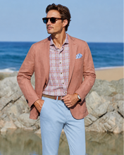 Man in a coral blazer and light blue pants standing by the ocean.