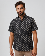 Man wearing a black patterned shirt with palm trees and beige pants on a white background